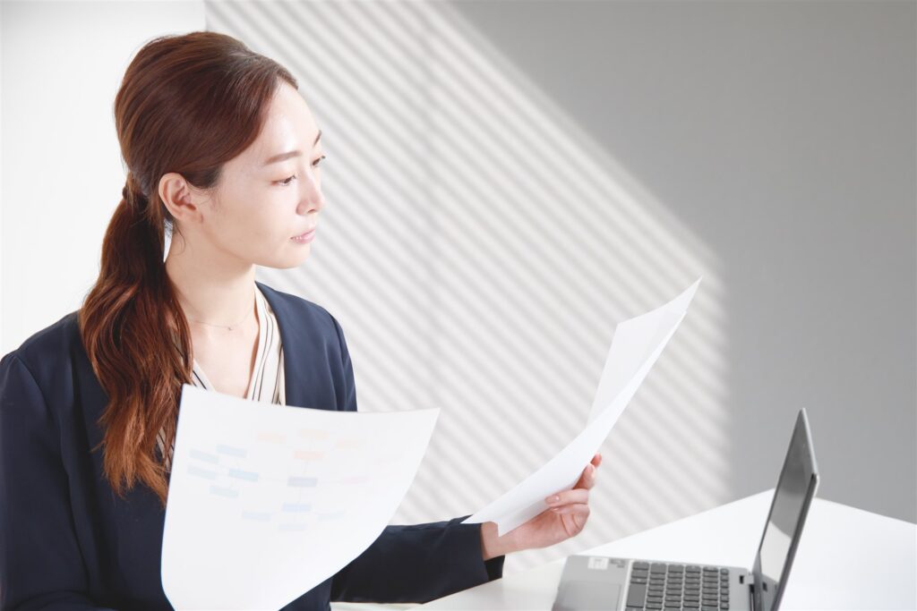書類を手にパソコンの前で考える女性