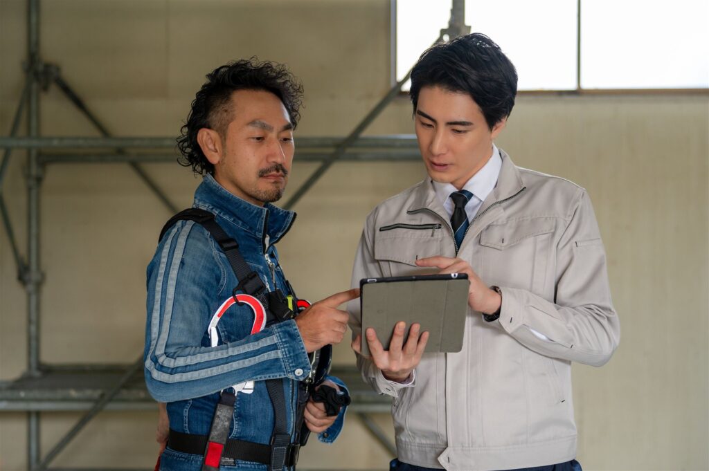 足場の前でタブレットを見て話す作業着の2人