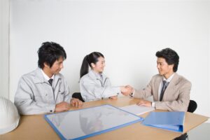 机をはさんで握手する作業着の2人と背広の男性