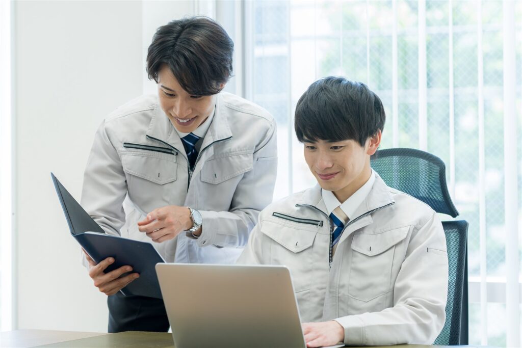 ノートパソコンと書類を前に相談する作業服の2人