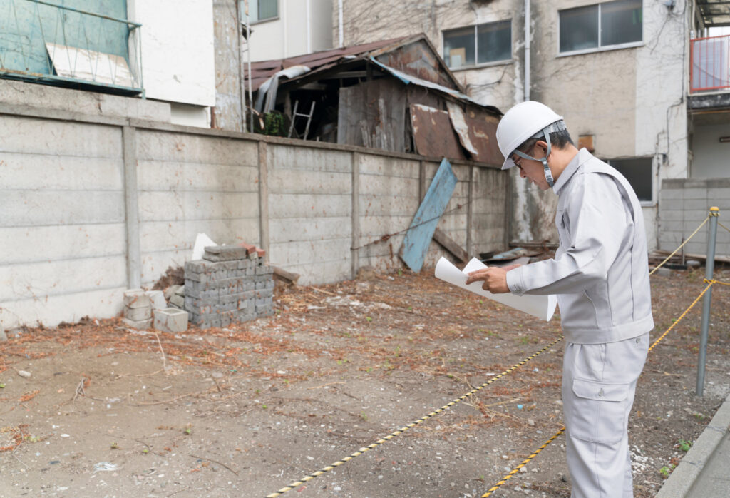 図面を持つ作業員が古い小屋の残る敷地を調べる様子