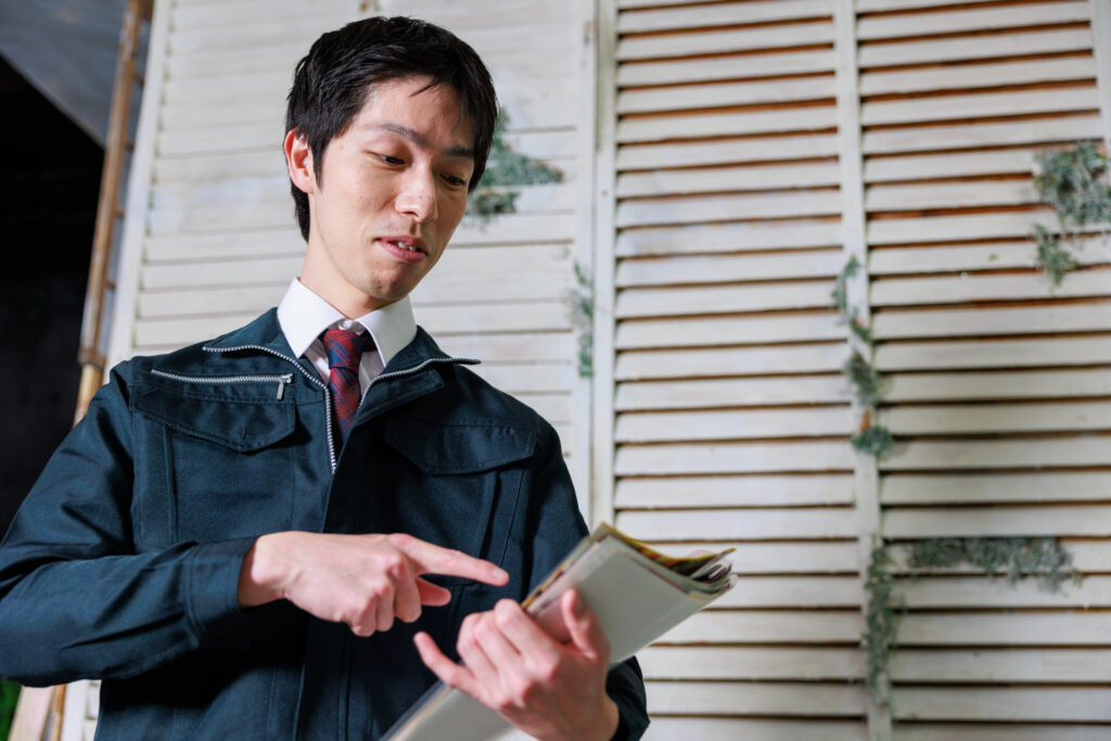 作業服にネクタイの男性が手元の冊子を指さして見直す