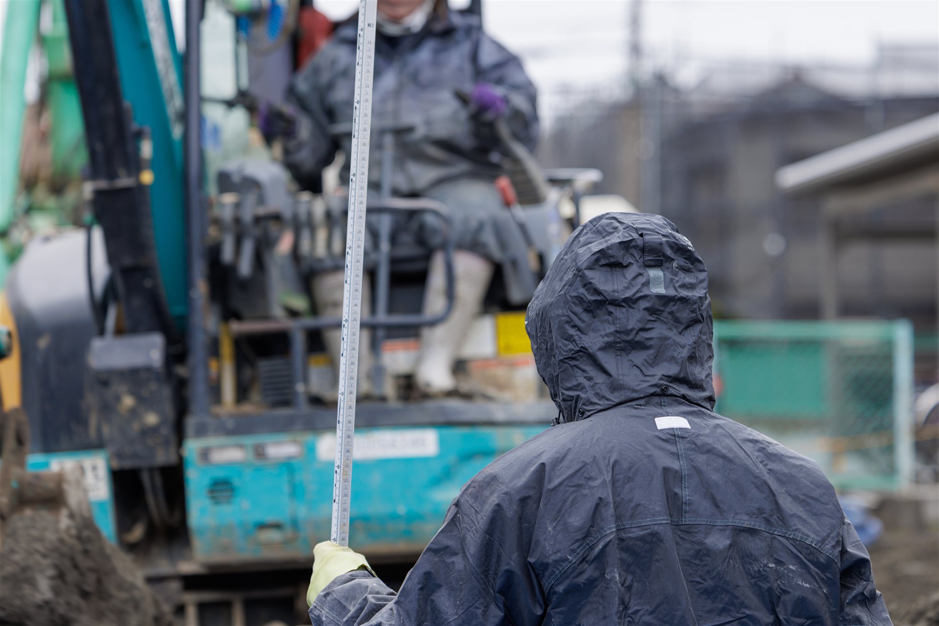 雨の中で雨がっぱを着て作業する人と重機（後ろ姿）