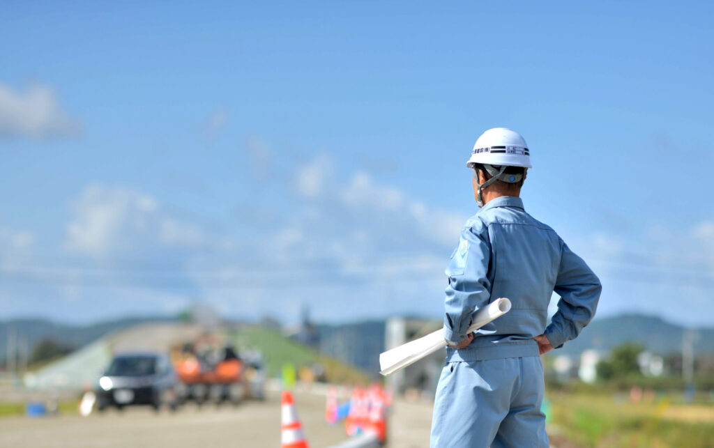 図面を持った作業着の監督が道路工事の現場を見渡す様子