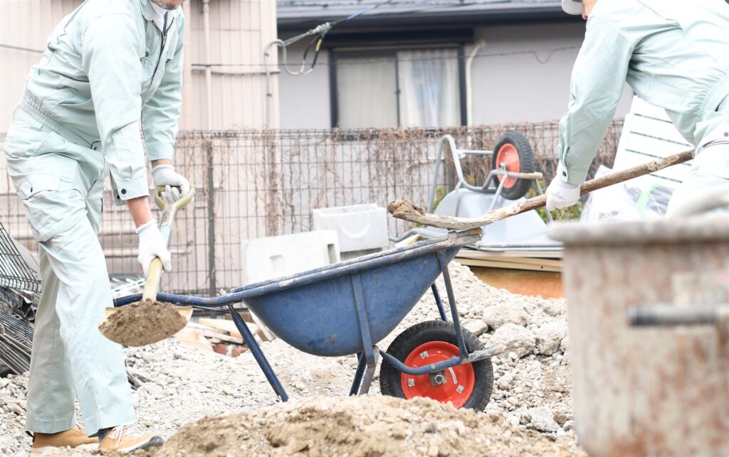 スコップで土を一輪車に入れる作業着の2人