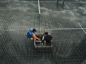 Two workers operating on a construction site with steel frameworks and concrete. High-angle view captures teamwork and industry.