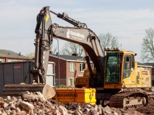 Yellow excavator working on a construction site, surrounded by rubble and buildings in the background.