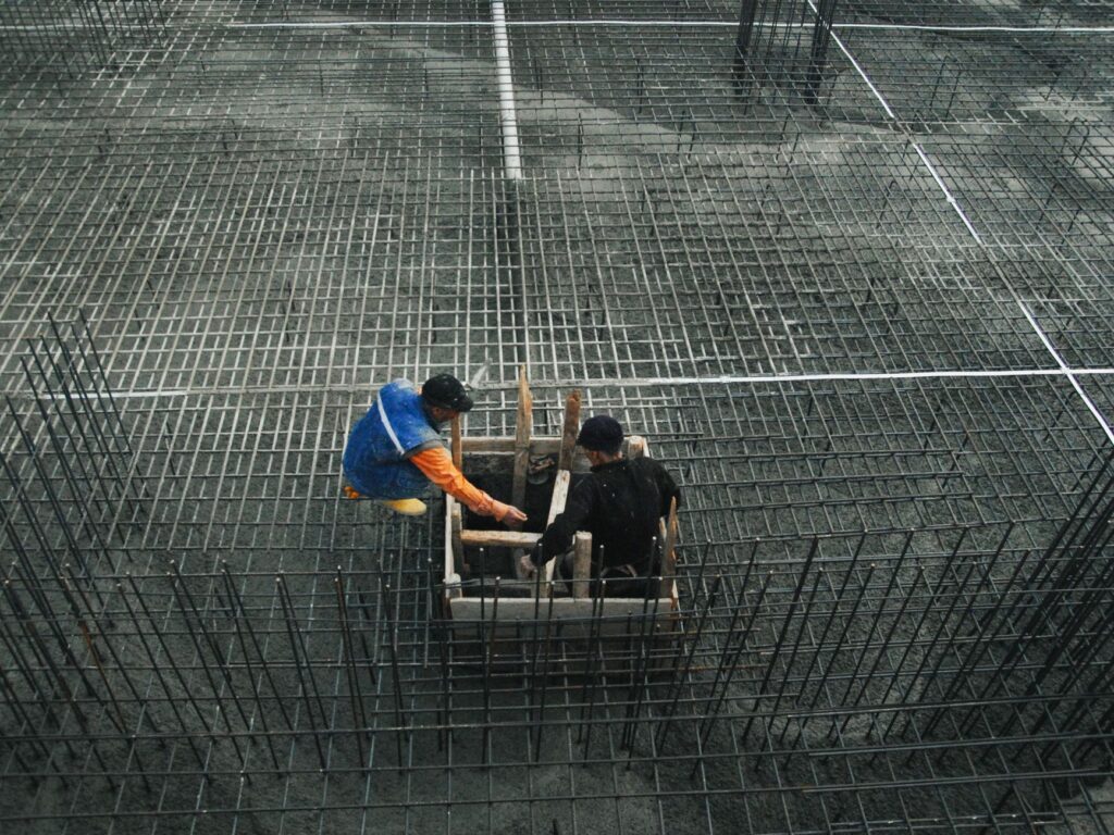 Two workers operating on a construction site with steel frameworks and concrete. High-angle view captures teamwork and industry.