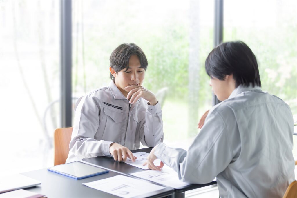 資料を見ながら打ち合わせる作業着の男性2人