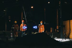 Workers in reflective gear and helmets conduct roadworks at night under bright lights.