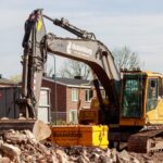 Yellow excavator working on a construction site, surrounded by rubble and buildings in the background.