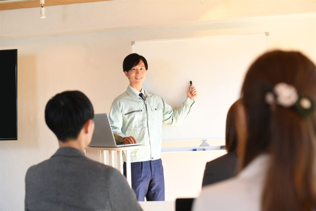 ホワイトボードの前で説明する作業着の男性と聞く人たち
