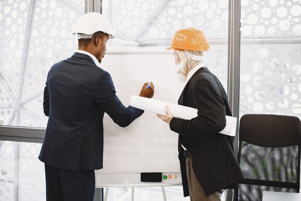 Two architects in hard hats working on a blueprint indoors, highlighting teamwork.