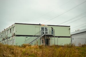 Exterior view of a green industrial building with stairs and fence under cloudy skies.