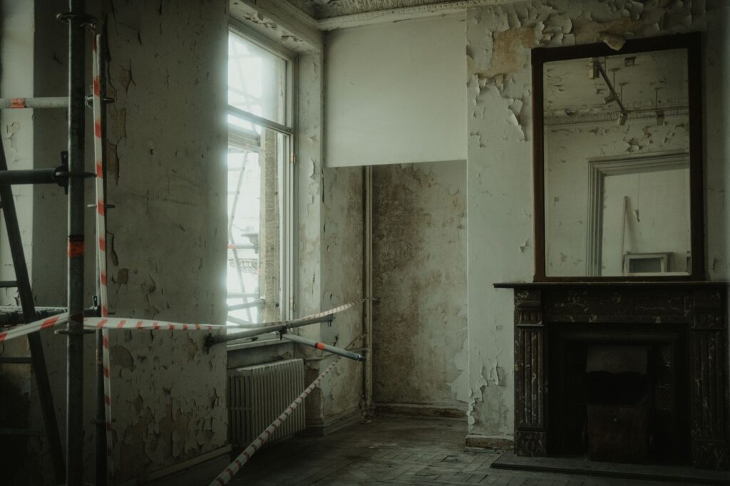 Abandoned room with peeling walls, construction tape, and a large mirror creating a creepy atmosphere.