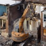 An excavator demolishes an old brick building at a construction site, creating debris and dust.