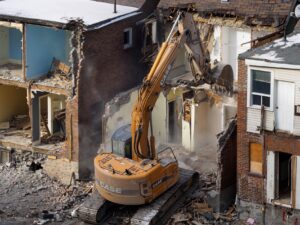 An excavator demolishes an old brick building at a construction site, creating debris and dust.