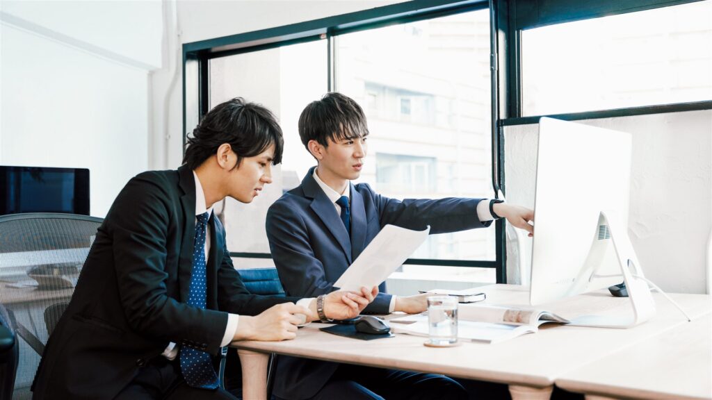 書類を手にモニターを指して確かめ合うスーツの男性2人