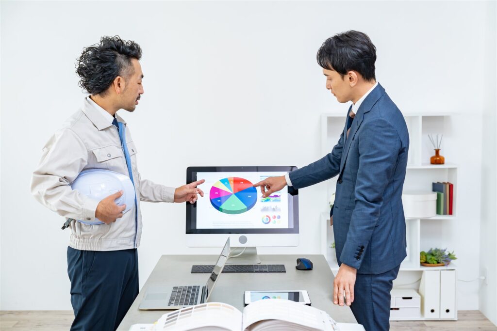 作業着の男性とスーツの男性が画面の円グラフで打ち合わせ