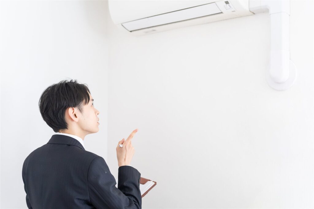 手帳を持ったスーツの男性が壁のエアコンを指さす様子