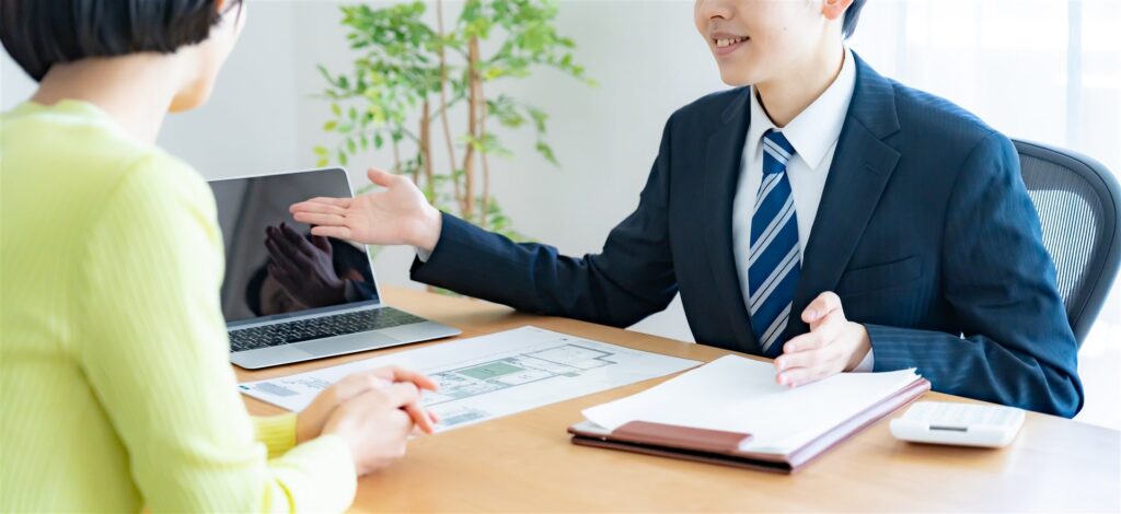机の図面を前にスーツの担当者が女性に説明する様子
