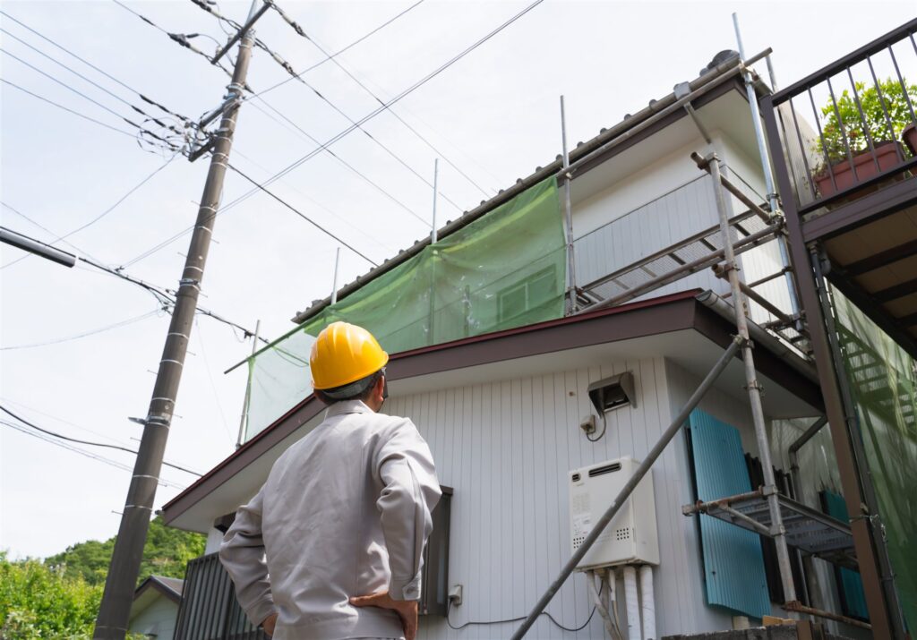 足場と養生の網が掛かった住宅を見上げる作業員の後ろ姿