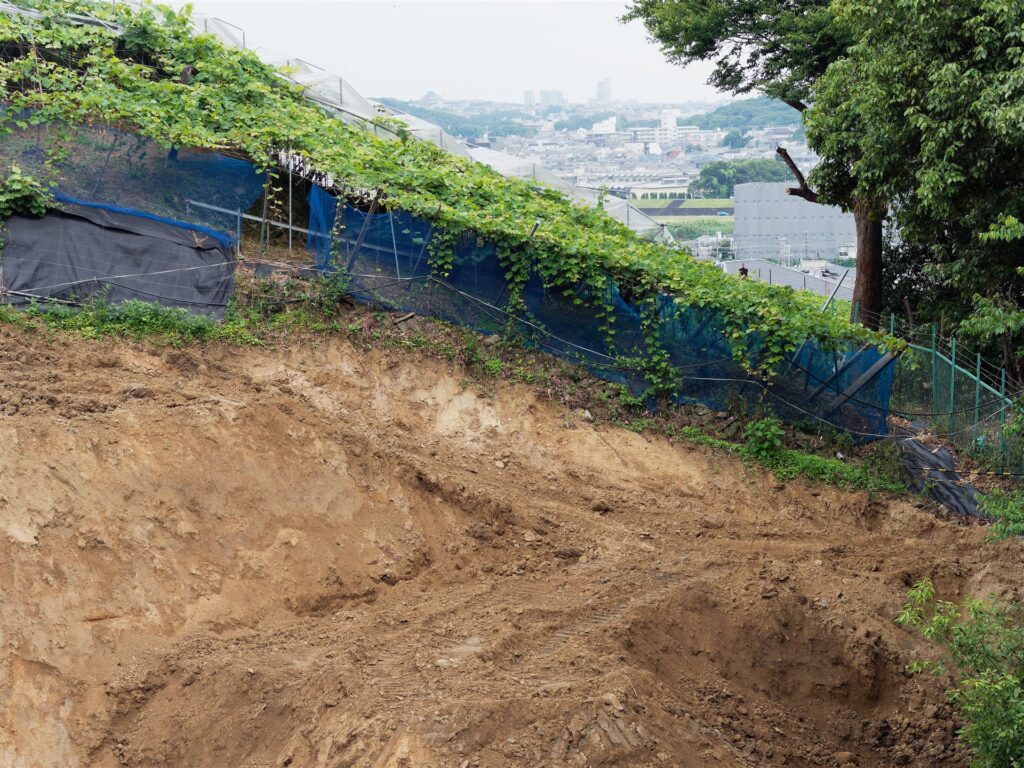 住宅地を見下ろす高台の削った土の斜面と青いネット
