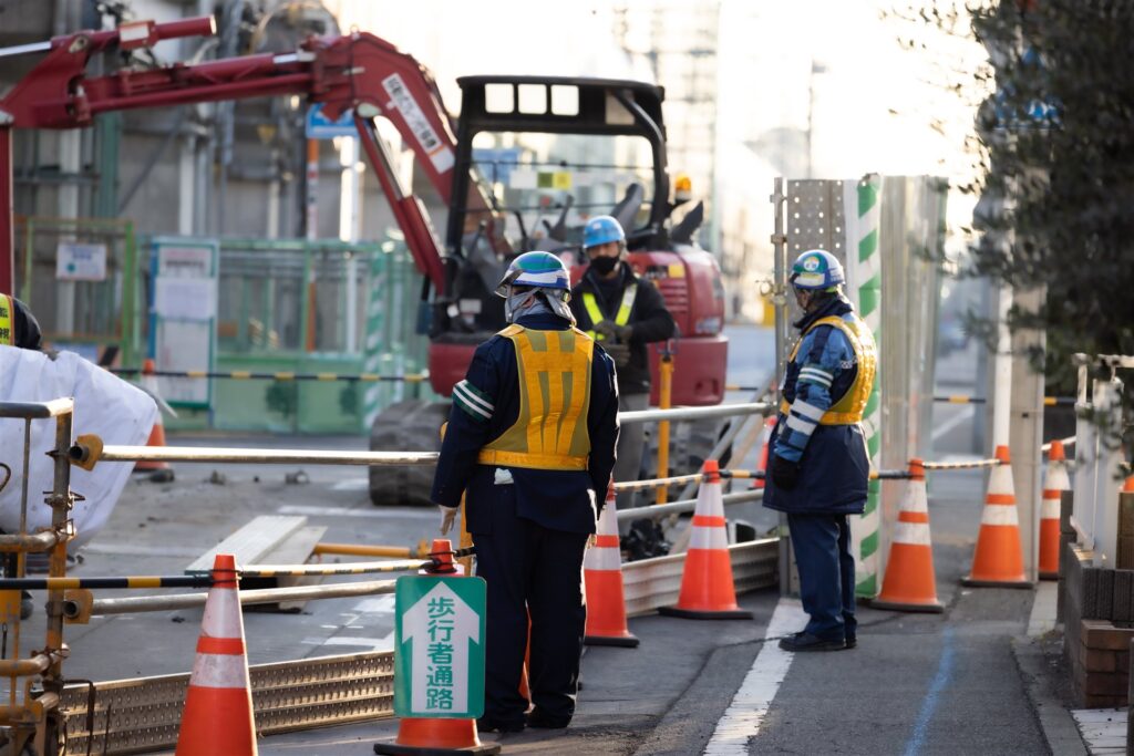 歩行者通路の札とコーンの間に立つ誘導員と小型のショベル