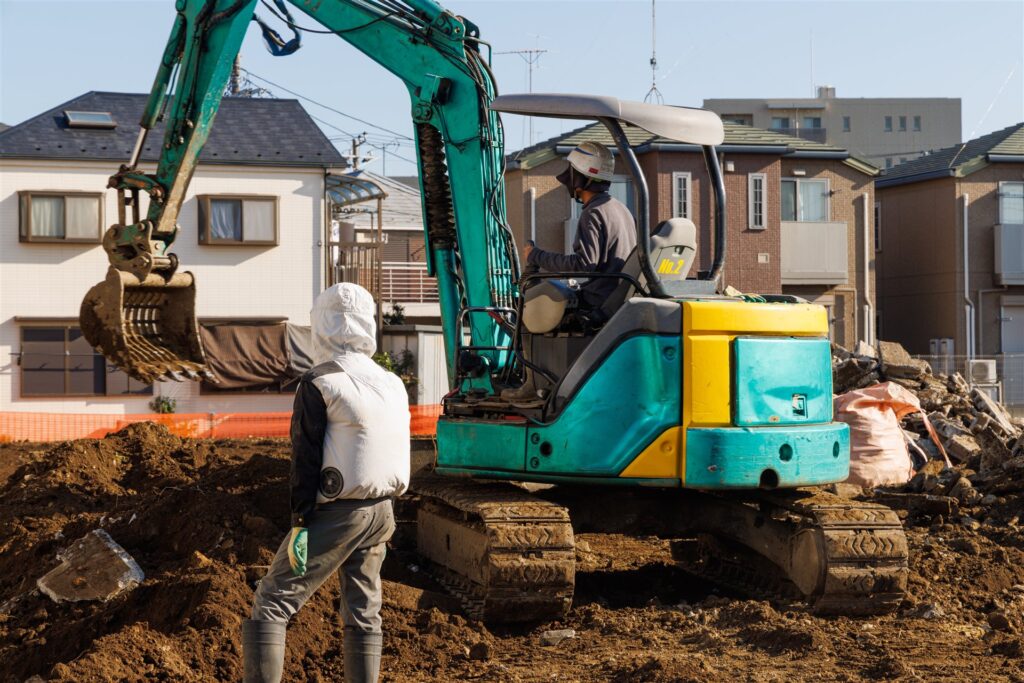住宅地の更地で小型の重機が土を掘り作業員が見守る